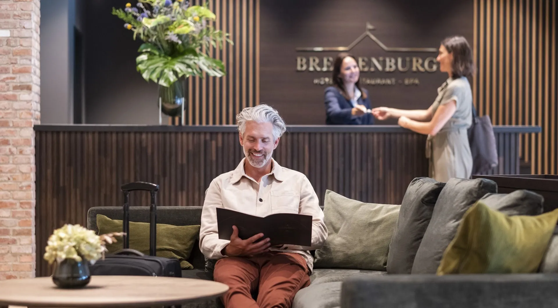 Mann sitzt im Wartebereich des Hotels, während Frau an der Rezeption steht.