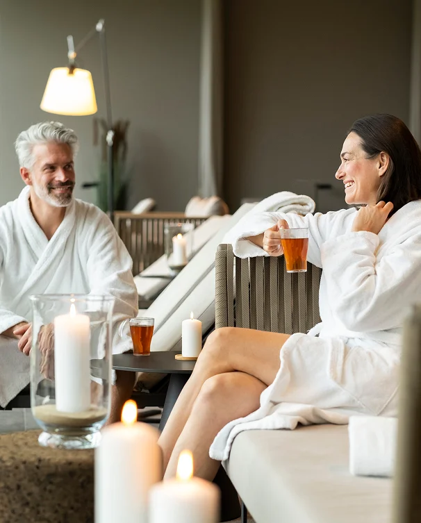 A couple in white bathrobes relaxing with tea in a spa lounge surrounded by candlelight.